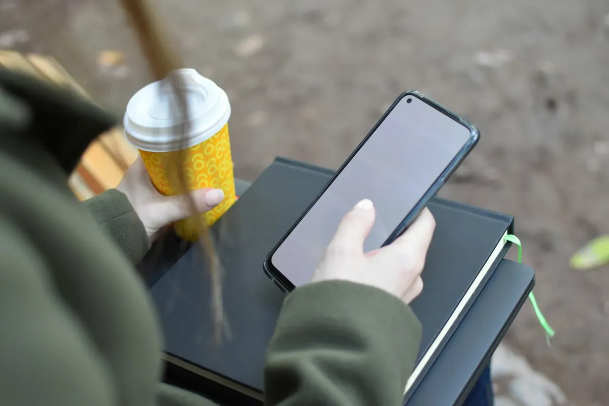 Une main qui tient un téléphone, un carnet et un café, dehors